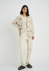 Beige linen button-up jacket with pockets, paired with a beige tank top and light-colored pants featuring contrast stitching and black slides.