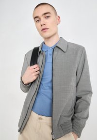 Sivý špicatý kabátik s golierovým dizajnom a zipsom, skombinovaný s modrým polo tričkom. Model drží popruh čierneho batohu.