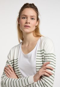 Bílý cardigan se zelenými horizontálními pruhy, s měkkou texturou a otevřeným designem vpředu. Nosený přes bílou košili, s překříženýma rukama.