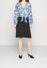 Top blu e bianco a fantasia con un nodo in vita, abbinato a una gonna nera. La modella indossa mule neri con punta affusolata.