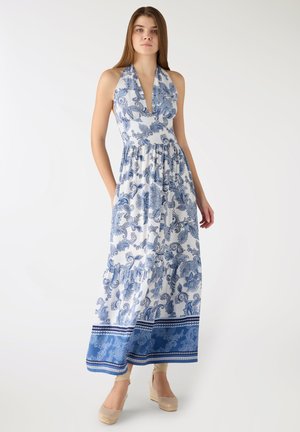 Giovane donna in piedi che indossa un vestito maxi bianco con spalline ad anello, decorato con motivi floreali blu, e sandali con zeppa beige.