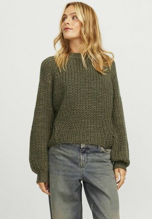 Gebreide trui in donkergroen met een gestructureerd, chunky patroon. Bevat een hoge halslijn en lange, geribbelde mouwen. Gecombineerd met lichtblauwe jeans.