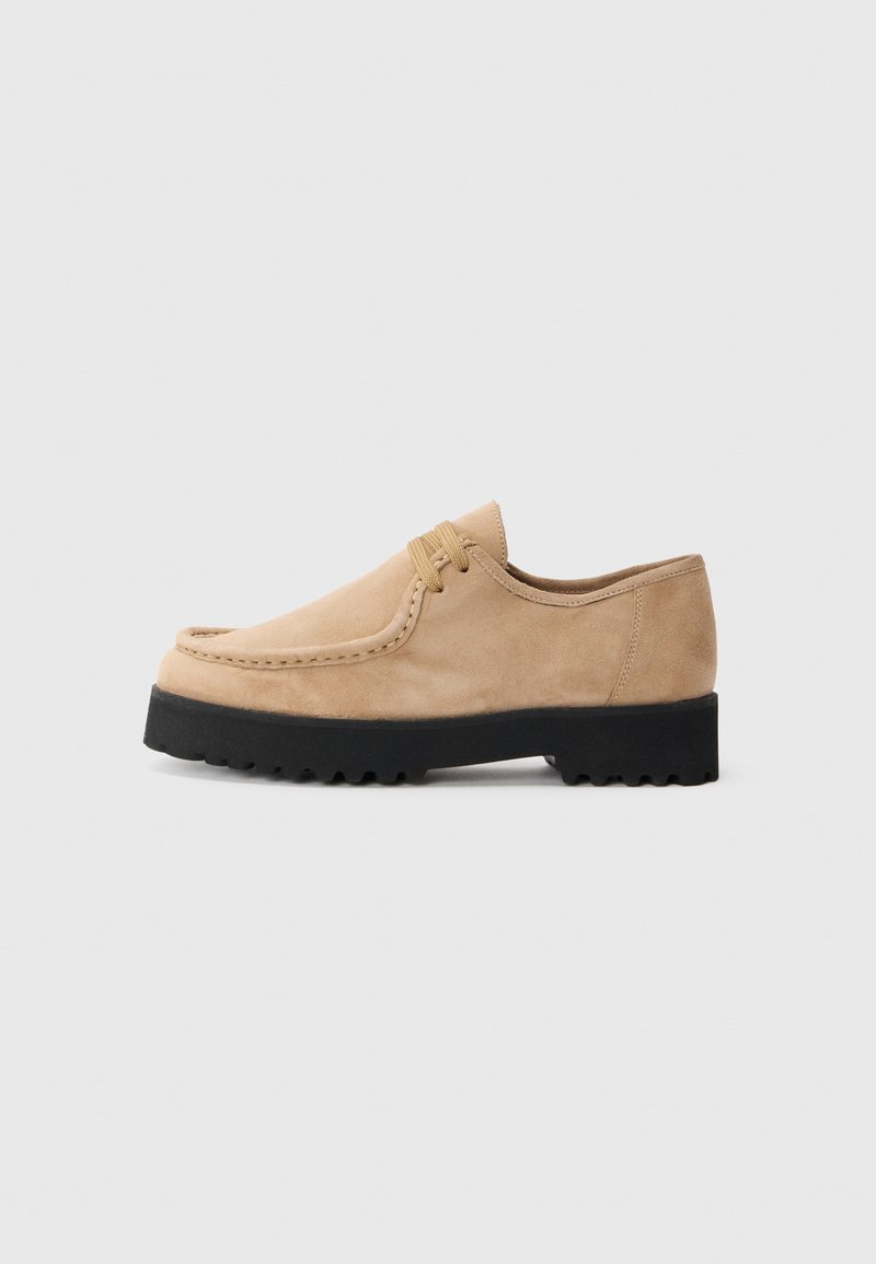 Beige semsket sko med rund tå, sydd detaljer og en svart gummisåle med mønstre for grep. Snøring på overdelen.