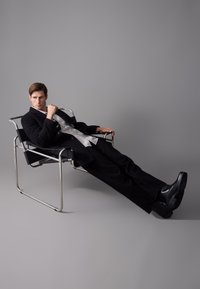 Silla de tela negra con estructura de cromo; modelo masculino sentado en abrigo negro y zapatos, reclinándose hacia atrás con una pierna extendida, fondo gris.