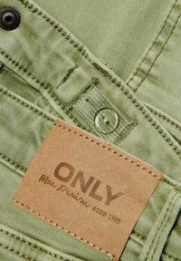 Tela de denim verde oliva con una superficie texturizada, costuras en contraste, un botón de metal y una etiqueta de cuero marrón con texto en relieve.