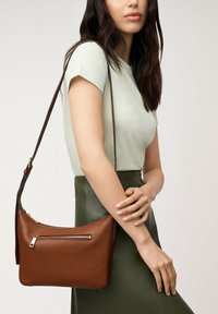 Sac en cuir marron à bandoulière avec une finition texturée, présentant une poche zippée à l'avant et une longue sangle sombre, exposé sur un fond neutre.