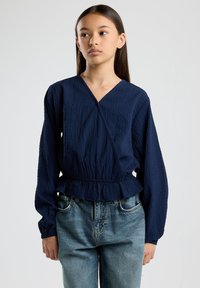 Navy blue wrap blouse with a textured finish, long sleeves, and a cinched waist, paired with light blue jeans featuring a classic cut.