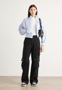 Giacca bomber blu chiaro con accenti neri, abbinata a pantaloni cargo neri con multiple tasche e cerniere, e sneakers nere.