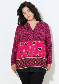 Vrouw met donker, krullend haar die een helderroze tuniek met sterren, harten en geometrische ontwerpen draagt, staande tegen een witte achtergrond.