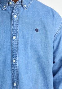 Lichtblauwe denim overhemd met button-down kraag, witte knopen en een klein marineblauw logo-embleem op de linker borst. Zachte textuur en klassiek ontwerp.