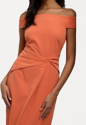 Kvinde iført en off-shoulder, rust-orange kjole med et foldet, knyttet detalje ved taljen og asymmetrisk drapering.