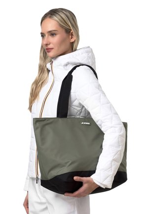 Donna bionda con giacca trapuntata bianca tiene sulla spalla una borsa a tracolla verde oliva e nera con manici neri.