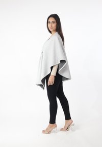 Poncho gris texturizado con diseño drapeado, cuello alto y abertura frontal. Combinado con leggings negros y sandalias transparentes de tacón grueso.