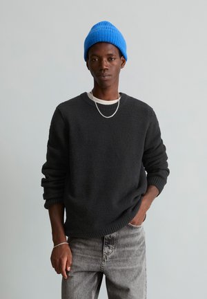 Jeune personne portant un bonnet bleu en tricot, un pull noir, un collier chaîne argenté, un jean gris, avec une main dans la poche sur un fond clair uni.