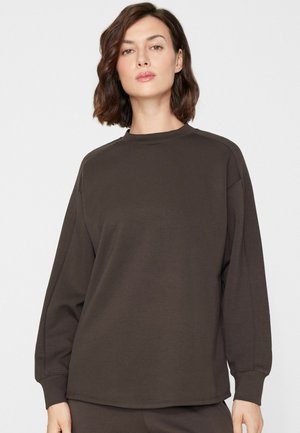 Femme aux cheveux courts et bruns portant un sweat-shirt ample à manches longues marron foncé sur un fond clair uni.