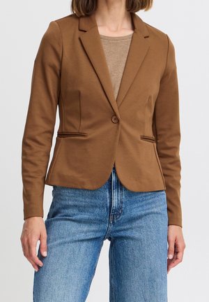 Femme portant un blazer marron ajusté sur un top beige associé à un jean bleu taille haute, sur un fond clair uni.