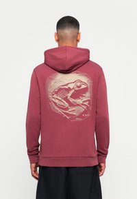 Bordeaux hettegenser med stor beinskjelett-frosk grafikk i beige på baksiden, med ribbet mansjetter og kant.