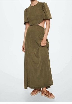 Femme portant une robe maxi vert olive à manches ballon, découpes sur les côtés et des sandales plates à lacets couleur beige tenant l'ourlet de la robe.