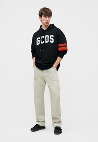 Ung mann iført en svart GCDS-hettegenser med røde striper på ermene, beige bukser med store lommer, og svarte joggesko, stående med hendene i lommene.