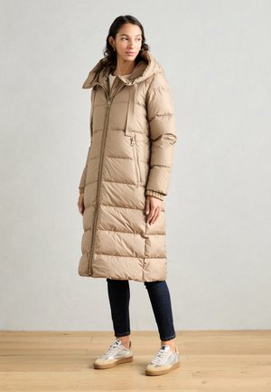 Beige puffer coat with a hood, long length, quilted design, front zipper, side pockets, paired with dark jeans and silver sneakers.