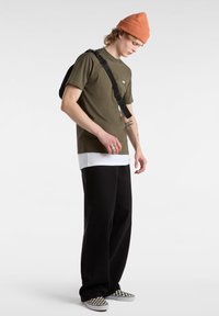 Camiseta verde oliva superpuesta sobre una camisa blanca, combinada con pantalones negros y zapatillas de cuadros. Gorro naranja y bolso negro de hombro.