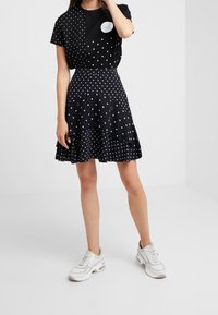 Chemise noire à pois avec des manches courtes et des pois blancs, associée à une jupe noire à pois superposée. Des baskets blanches complètent la tenue.