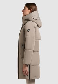 Puffer coat in light beige with a high collar, zip pockets, and a logo patch. Features a detachable hood and smooth, quilted texture.