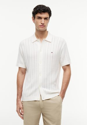 Mężczyzna ubrany w biały krótki swetrowy polo z pionowymi paskami i beżowe spodnie, stojący na jednolitym tle.