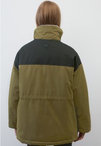 Grüne und schwarze gepolsterte Jacke mit hohem Kragen, die eine glatte Textur aufweist und ein Logo-Patch auf dem Rücken hat. Der Saum ist abgerundet.