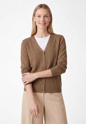 Smilende kvinde med lige blondt hår iført en brun knapbeklædt cardigan over en hvid skjorte og beige bukser, stående med den ene arm krydset.