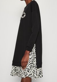 Schwarzes Sweatshirtkleid mit langen Ärmeln, das einen gepunkteten Saum aus Leopardenmuster in Weiß und Schwarz aufweist. Logo-Detail auf der Vorderseite. Aus weichem Baumwollmix.