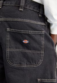 Pantalones cargo de mezclilla negra con costuras en contraste, que presentan un bolsillo parche en la parte trasera con una etiqueta de logo "Dickies" multicolor.