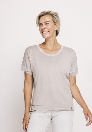 Smiling woman with short blonde hair wears a light beige short-sleeve top and white pants, standing against a plain white background.