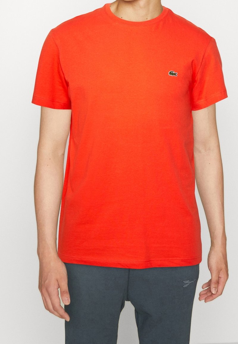 Personne portant un t-shirt à manches courtes orange vif avec un petit logo de crocodile vert sur la poitrine et un pantalon gris foncé.