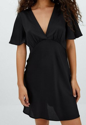 Femme portant une courte robe noire à manches flottantes et un décolleté en V profond, debout les bras détendus le long du corps.