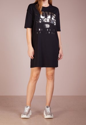 Mustat ylisuurten T-paitamekot, joissa on metallinhohtoinen "LOVE MOSCHINO" -painatus. Lyhyet hiharat, pyöreä pääntie ja sileä kangas. Yhdistetty harmaisiin sneaker-kengiin.