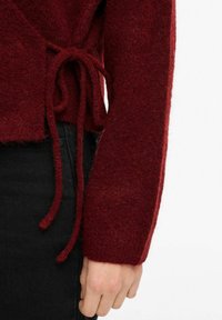 Bordo vilnos megztinis su tekstūruotu paviršiumi, ilgomis rankovėmis ir surišimu ties liemeniu, derinamas su juodais džinsais.