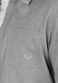 Poche de chemise en tissu gris avec une texture diagonale subtile et un petit logo brodé de deux feuilles à l'avant.