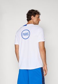 T-shirt de sport blanc à manches courtes, avec un empiècement bleu sur l'épaule et un logo circulaire "PARIS" dans le dos.