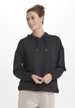 Sweatshirt à capuche noir avec une coupe décontractée, doté d'un cordon de serrage et de poignets élastiques, fabriqué en tissu doux. Porté sur un pantalon beige clair.