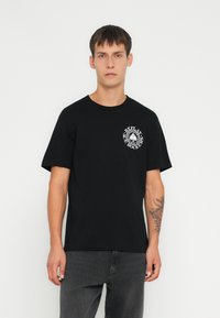 Camiseta de algodón negra con mangas cortas, escote redondo y logotipo gráfico blanco en el lado izquierdo del pecho. Ajuste casual, textura suave.