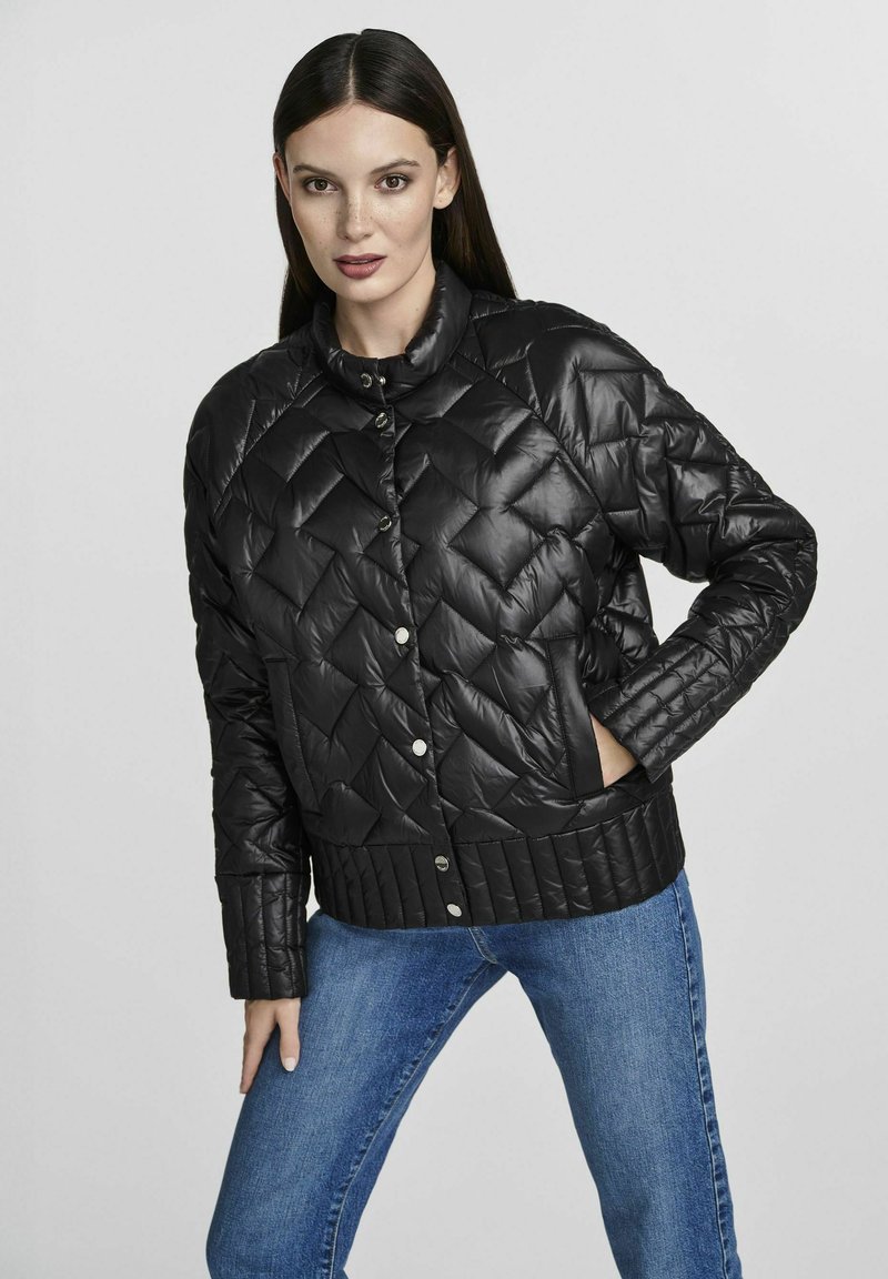 Giacca nera imbottita con motivo a rombo, chiusura a bottoni a pressione, fondo a costine e tasche laterali. Indossata con jeans blu.