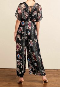 Combinaison noire à fleurs avec des manches courtes à volants, présentant des motifs complexes rouges et bleus et un détail en keyhole au dos. Design à jambes larges.
