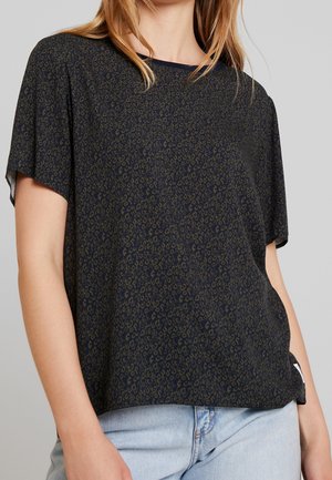 Woman wearing a dark short-sleeve leopard print top and light blue jeans, shown from shoulders to hips against a plain background.