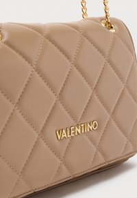 Prešívaná béžová kabelka s pozlátenou kovovou výbavou, so značkovým detailom a retiazkovým popruhom. Hladká textúra s diamantovým vzorom.