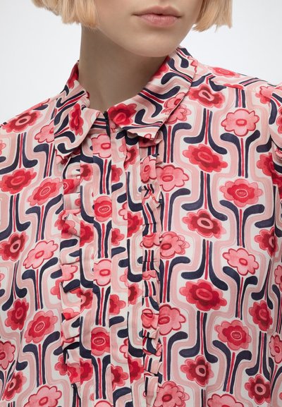 Femme portant un chemisier rose avec un motif floral rouge et bleu marine et des lignes ondulées, avec un devant à volants et un col pointu.