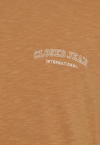 Bruine stof met witte geborduurde tekst "CLOSED JEAN INTERNATIONAL" in een gebogen en rechte indeling.