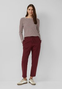 Vrouw die staat met handen in haar zakken, gekleed in een bordeauxrode en witte gestreepte lange mouw top, bordeauxrode broek en witte sneakers op een grijze achtergrond.