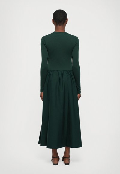 Long-sleeve dress in deep green fabric, featuring a fitted bodice and a gathered waist, flowing into a full-length skirt.