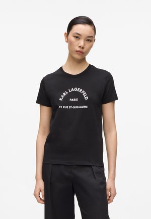 Femme aux cheveux foncés portant un t-shirt noir Karl Lagerfeld avec un texte blanc, associé à un pantalon noir, se tenant devant un fond clair uni.